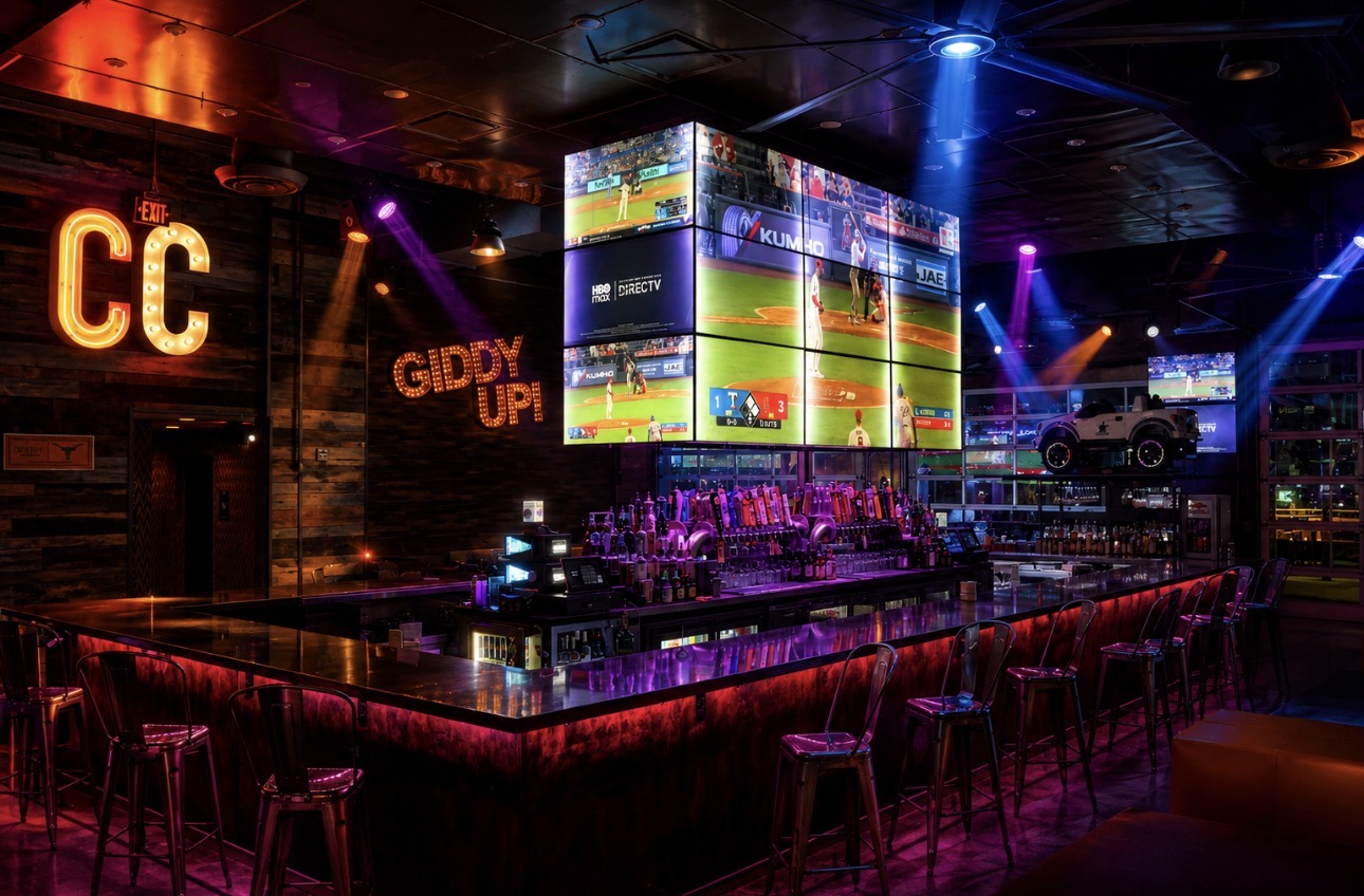 Concrete Cowboy Frisco interior with CC marquee sign, GIDDY UP neon, and a wall of sports screens behind the bar
